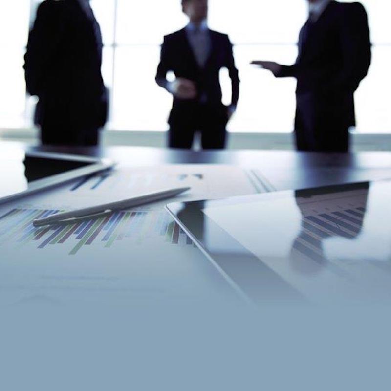 A tablet, pen and documents with charts on a table. Three business men in the background are blurred.